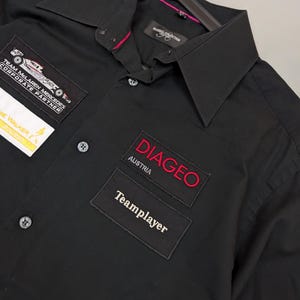 Puede incluir: Camisa negra con botones y parches. Los parches incluyen "Team McLaren Mercedes Corporate Partner", "DIAGEO AUSTRIA" y "Teamplayer". La camisa tiene cuello y est&aacute; colgada de una percha negra.