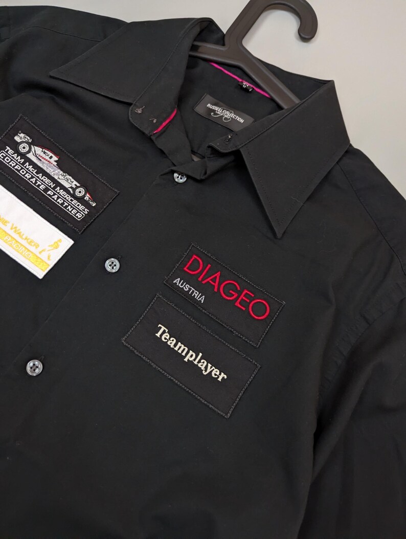 Puede incluir: Camisa negra con botones y parches. Los parches incluyen "Team McLaren Mercedes Corporate Partner", "DIAGEO AUSTRIA" y "Teamplayer". La camisa tiene cuello y est&aacute; colgada de una percha negra.