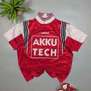 May include: A red Adidas Akku Tech jersey with white sleeves and a white collar with black and red stripes. The jersey has the Adidas logo and the text "AKKU TECH" in a white box.