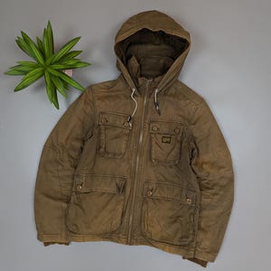 May include: Olive green hooded jacket with multiple front pockets and a zipper closure. The jacket features a drawstring hood and a small logo patch. The jacket is made of a durable, possibly waxed, fabric.