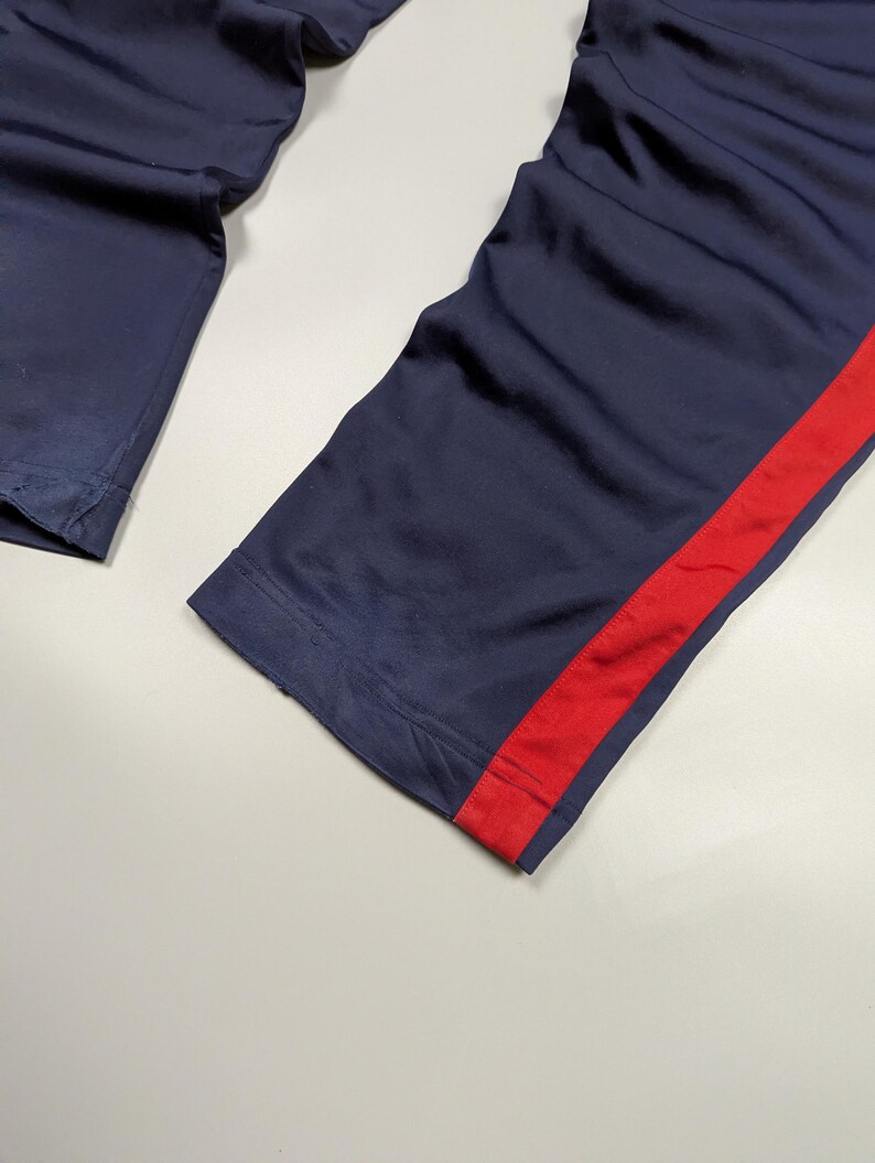 May include: Navy blue athletic trousers with a bold red stripe along the side. The fabric looks smooth, possibly synthetic. The image is a close-up of the lower part of the trousers.