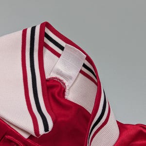 May include: Close-up of a red athletic jersey with a white collar. The collar has black and red stripes. A white tag is sewn into the collar.