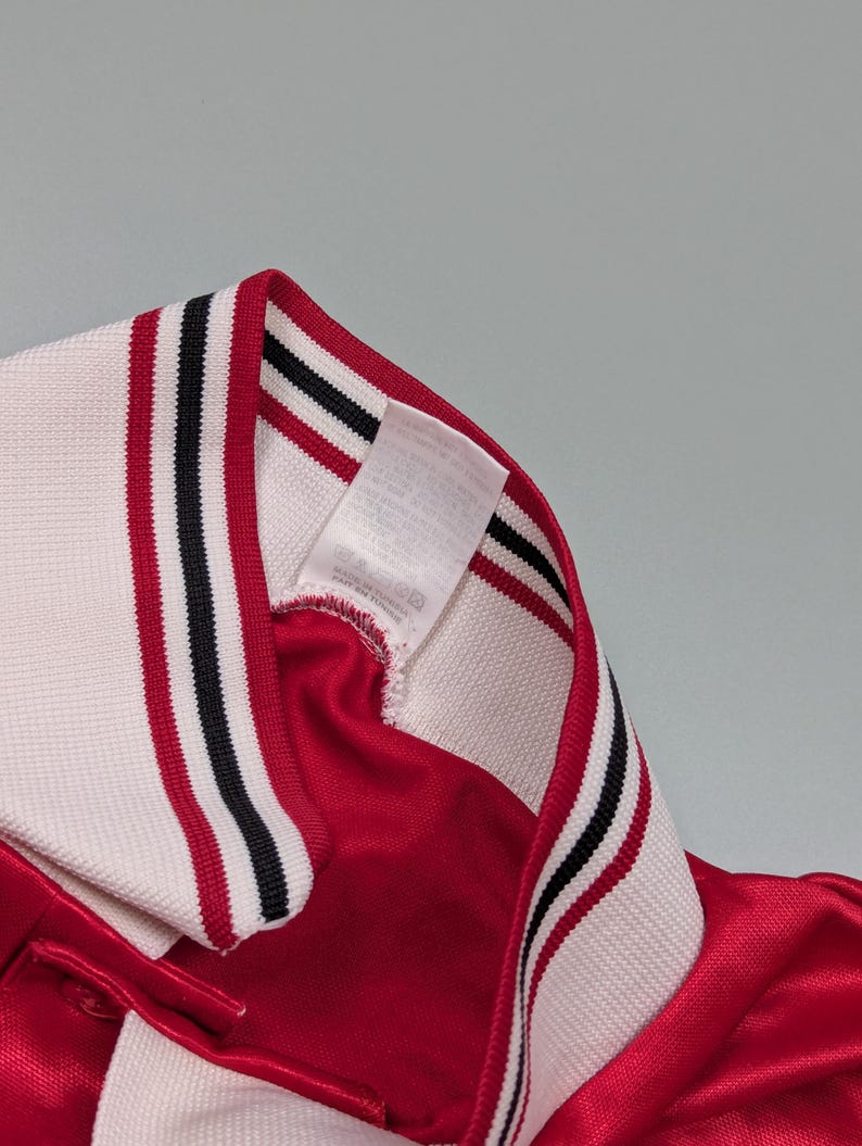 May include: Close-up of a red athletic jersey with a white collar. The collar has black and red stripes. A white tag is sewn into the collar.