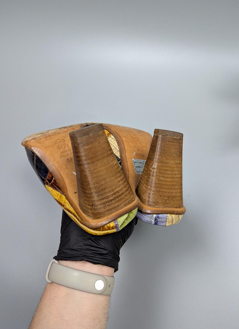 May include: A pair of colorful heeled shoes with a patterned design. The heels are a light brown color and the soles are visible. The shoes are being held up by a person wearing a black glove.