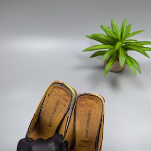 May include: A pair of Gianmarco Lorenzi heeled mules. The shoes feature a colorful pattern with shades of yellow, purple, and pink. The interior is a light brown color with the brand name printed on the insole. A black gloved hand holds the shoes.