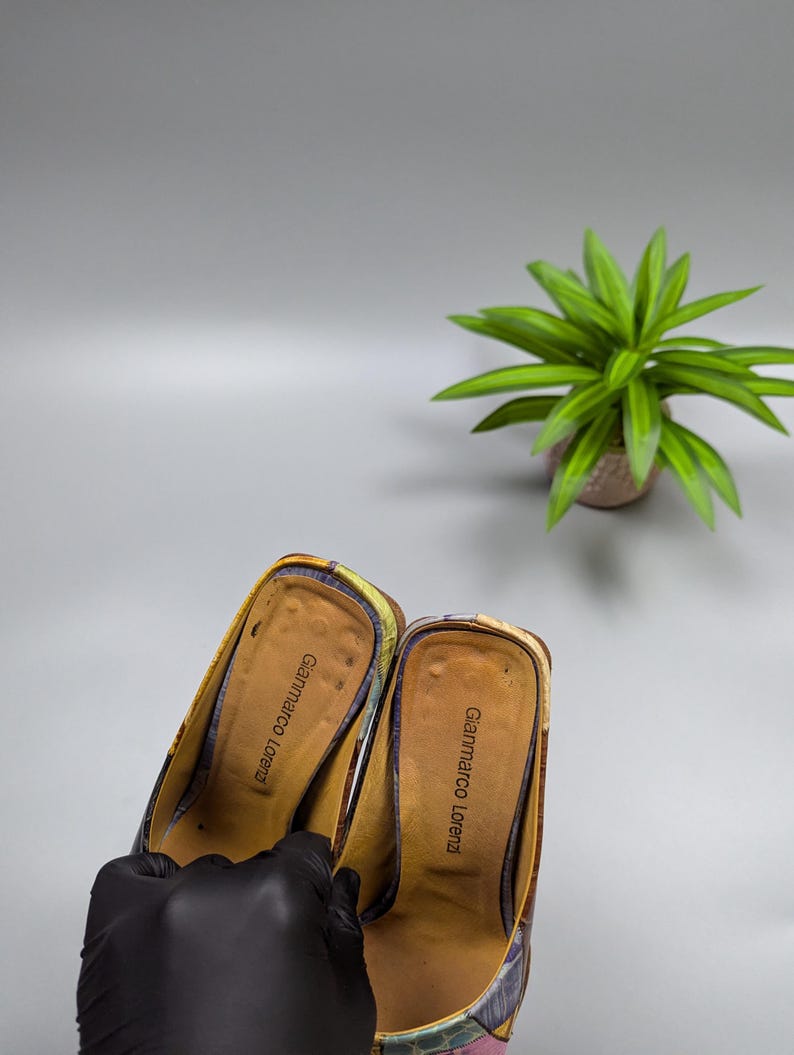 May include: A pair of Gianmarco Lorenzi heeled mules. The shoes feature a colorful pattern with shades of yellow, purple, and pink. The interior is a light brown color with the brand name printed on the insole. A black gloved hand holds the shoes.