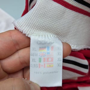 May include: Close-up of a white tag with the Adidas logo and size information. The tag is attached to a red and white striped collar. The tag also includes flags and the text "100% polyester".