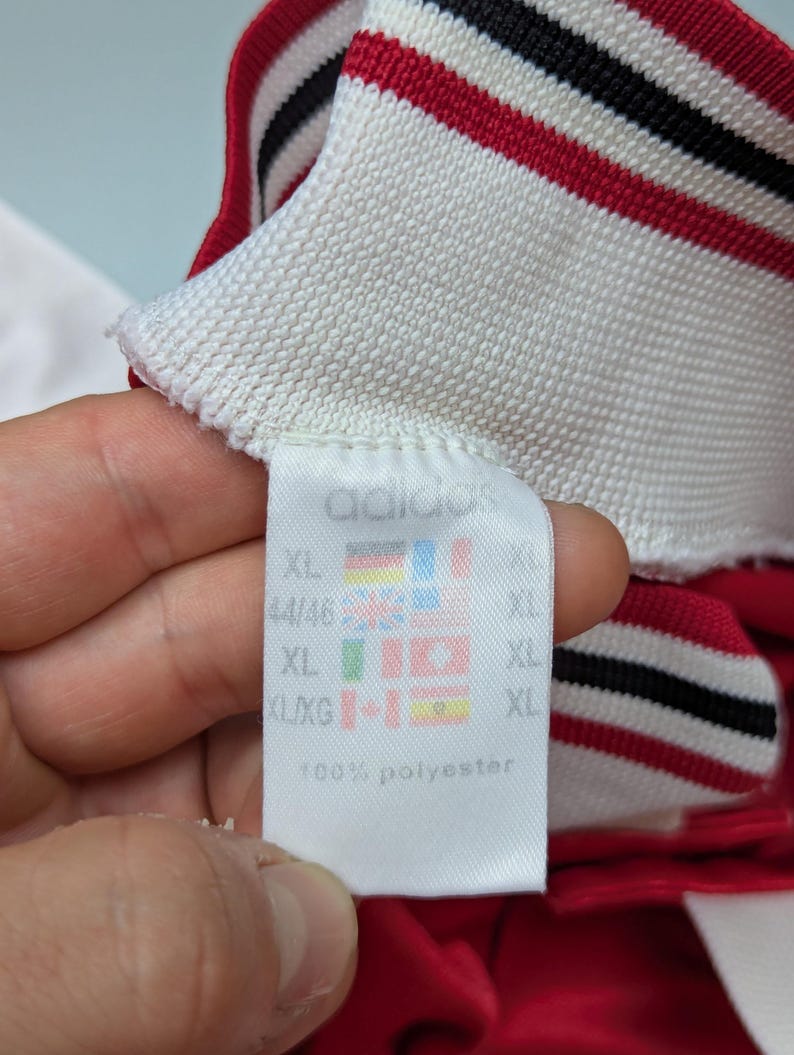 May include: Close-up of a white tag with the Adidas logo and size information. The tag is attached to a red and white striped collar. The tag also includes flags and the text "100% polyester".