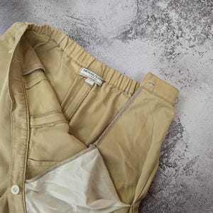 May include: A pair of tan trousers featuring an elasticized waistband, a side button closure, and a zipper fly. The interior label reads Michael Glasser. The garment is constructed from a solid-colored twill fabric with a visible white lining.