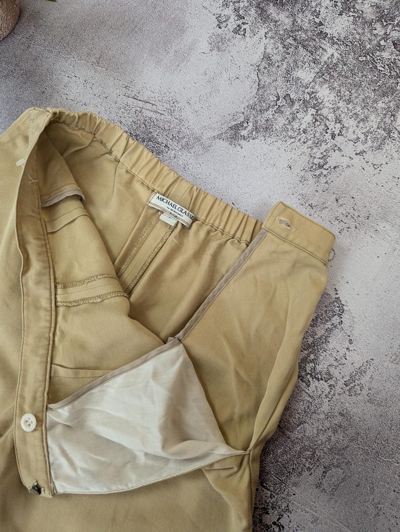 May include: A pair of tan trousers featuring an elasticized waistband, a side button closure, and a zipper fly. The interior label reads Michael Glasser. The garment is constructed from a solid-colored twill fabric with a visible white lining.