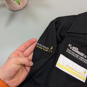 Puede incluir: Camisa negra con parches bordados. Los parches incluyen "WALKING JOHNNIE WALKER", "TEAM MCLAREN MERCEDES CORPORATE PARTNER" y "JOHNNIE WALKER RACING".