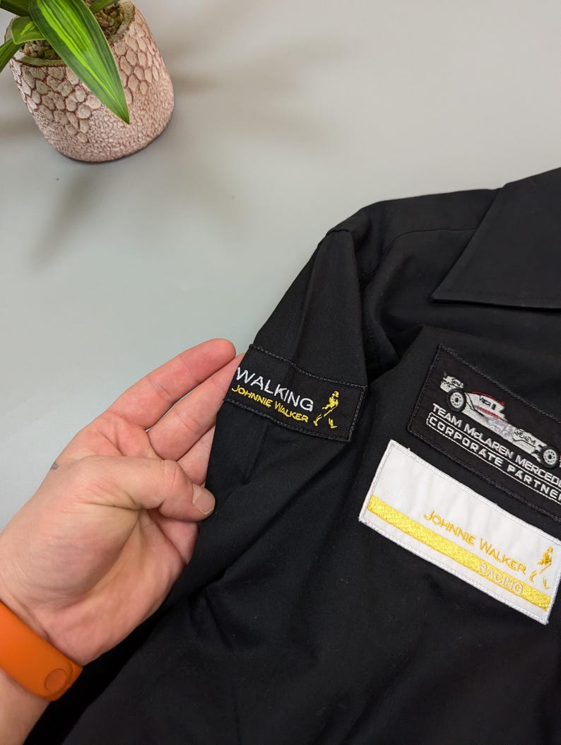 Puede incluir: Camisa negra con parches bordados. Los parches incluyen "WALKING JOHNNIE WALKER", "TEAM MCLAREN MERCEDES CORPORATE PARTNER" y "JOHNNIE WALKER RACING".