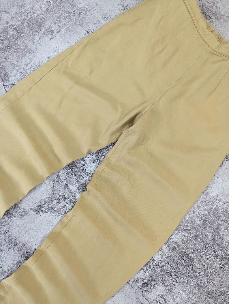 May include: A pair of solid light tan trousers featuring a high-waisted design with a visible elastic waistband at the top. The pants have a clean, straight-leg cut with subtle vertical seam detailing running down the front of each leg.
