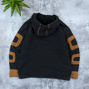 Pode incluir: Uma camisola com capuz preta apresentando pain&eacute;is de tecido castanho nas mangas e no capuz. A pe&ccedil;a inclui um capuz com detalhes de costura em castanho e mangas compridas com acentos castanhos sobre o corpo principal de cor preta.