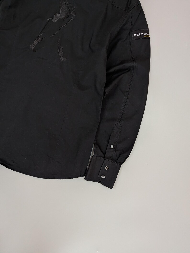 Puede incluir: Camisa negra de manga larga. Presenta un sutil dise&ntilde;o en relieve en la parte superior izquierda del pecho y un peque&ntilde;o parche rectangular en la manga derecha con el texto "KEEP WAL". La camisa tiene cuello cl&aacute;sico y pu&ntilde;os abotonados.