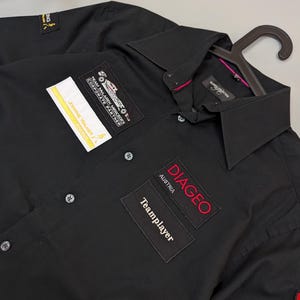 Puede incluir: Camisa negra con botones y varios parches. Los parches incluyen "Johnnie Walker", "Team McLaren Mercedes Corporate Partner" y "Diageo Austria Teamplayer". La camisa est&aacute; en una percha.