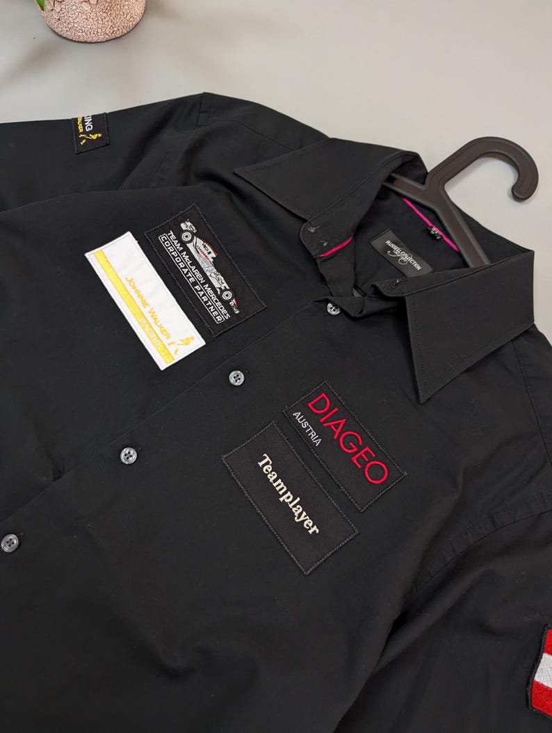 Puede incluir: Camisa negra con botones y varios parches. Los parches incluyen "Johnnie Walker", "Team McLaren Mercedes Corporate Partner" y "Diageo Austria Teamplayer". La camisa est&aacute; en una percha.