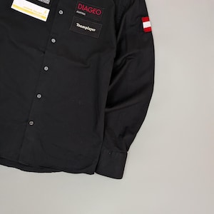 Puede incluir: Camisa negra abotonada de manga larga. Cuenta con parches con el texto "DIAGEO AUSTRIA" y "Teamplayer". Se incluye un peque&ntilde;o parche con la bandera austriaca y otro parche con texto. La camisa tiene cuello y pu&ntilde;os abotonados.
