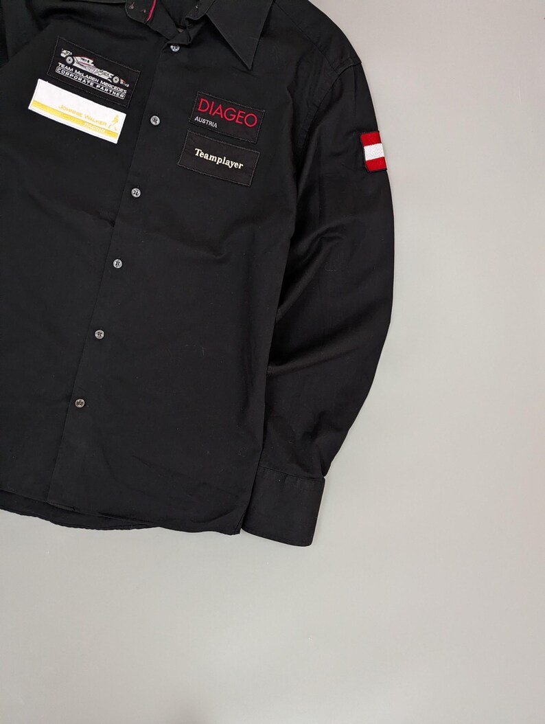 Puede incluir: Camisa negra abotonada de manga larga. Cuenta con parches con el texto "DIAGEO AUSTRIA" y "Teamplayer". Se incluye un peque&ntilde;o parche con la bandera austriaca y otro parche con texto. La camisa tiene cuello y pu&ntilde;os abotonados.