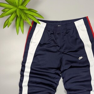 May include: Navy blue athletic trousers with white and red side stripes. The trousers feature the Nike logo in white on the front. The trousers are made of a shiny, smooth material.