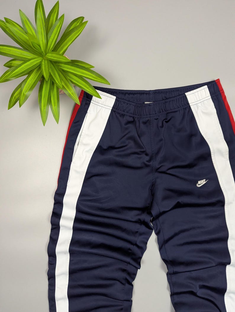 May include: Navy blue athletic trousers with white and red side stripes. The trousers feature the Nike logo in white on the front. The trousers are made of a shiny, smooth material.