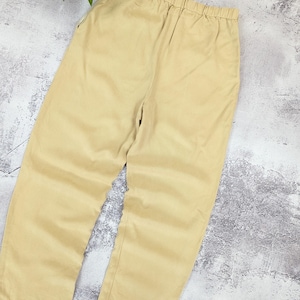 May include: A pair of light beige trousers featuring a comfortable elasticized waistband. These pants have a straight-leg cut and a simple, clean design with no visible pockets or embellishments, presented in a solid, neutral tan color.