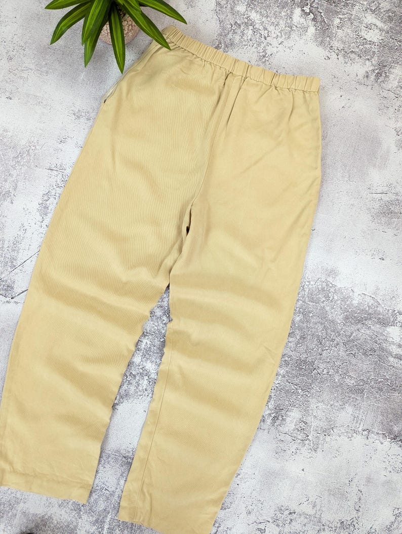 May include: A pair of light beige trousers featuring a comfortable elasticized waistband. These pants have a straight-leg cut and a simple, clean design with no visible pockets or embellishments, presented in a solid, neutral tan color.