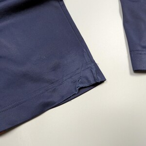 May include: Close-up of navy blue fabric, possibly part of a garment. The fabric has a smooth texture and appears to be a medium weight material. The edge of the fabric is visible, showing a seam and a slight fraying. The overall impression is of a tailored piece.
