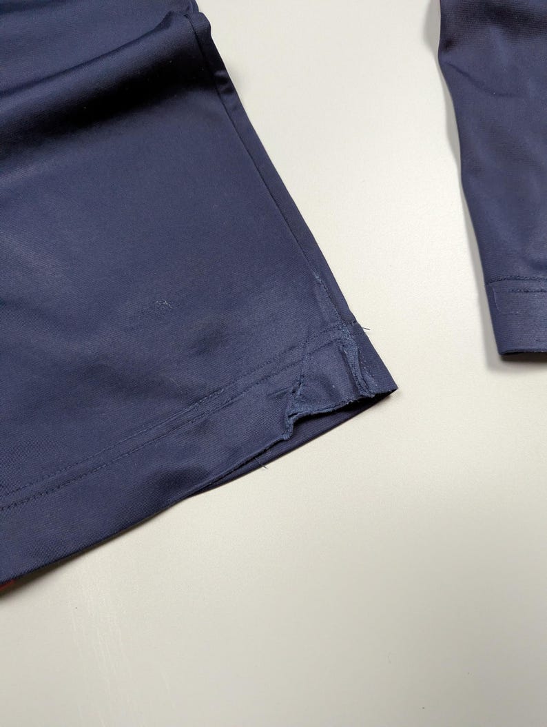 May include: Close-up of navy blue fabric, possibly part of a garment. The fabric has a smooth texture and appears to be a medium weight material. The edge of the fabric is visible, showing a seam and a slight fraying. The overall impression is of a tailored piece.