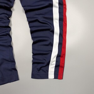 May include: Navy blue tracksuit bottoms with a white stripe and a red stripe down the side. The trousers appear to be made of a smooth, synthetic material. The image shows the lower part of the trousers.