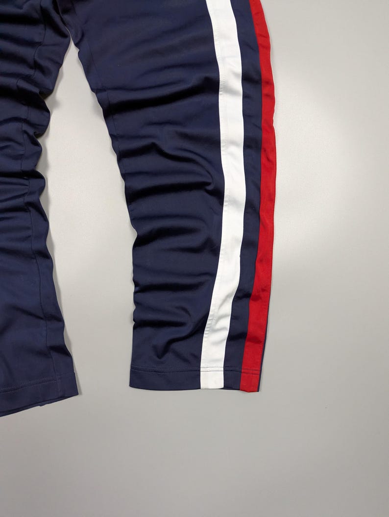 May include: Navy blue tracksuit bottoms with a white stripe and a red stripe down the side. The trousers appear to be made of a smooth, synthetic material. The image shows the lower part of the trousers.