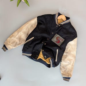 Pode incluir: Casaco universitário preto e creme com mangas de couro. O casaco apresenta um patch com a letra "S" e um forro dourado. A gola e os punhos têm um padrão azul marinho e branco.