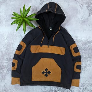 Pode incluir: Um casaco preto com capuz, fecho de meio z&iacute;per e cord&atilde;o. A pe&ccedil;a apresenta pain&eacute;is em tom castanho nas mangas, um bolso frontal grande e uma aba no peito. Um emblema em forma de cruz preta est&aacute; centralizado no bolso da frente.