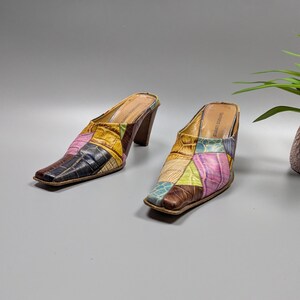 May include: A pair of patchwork leather mules with a square toe and a brown heel. The mules feature a variety of colors, including pink, blue, yellow, and brown. The shoes are set against a gray background.