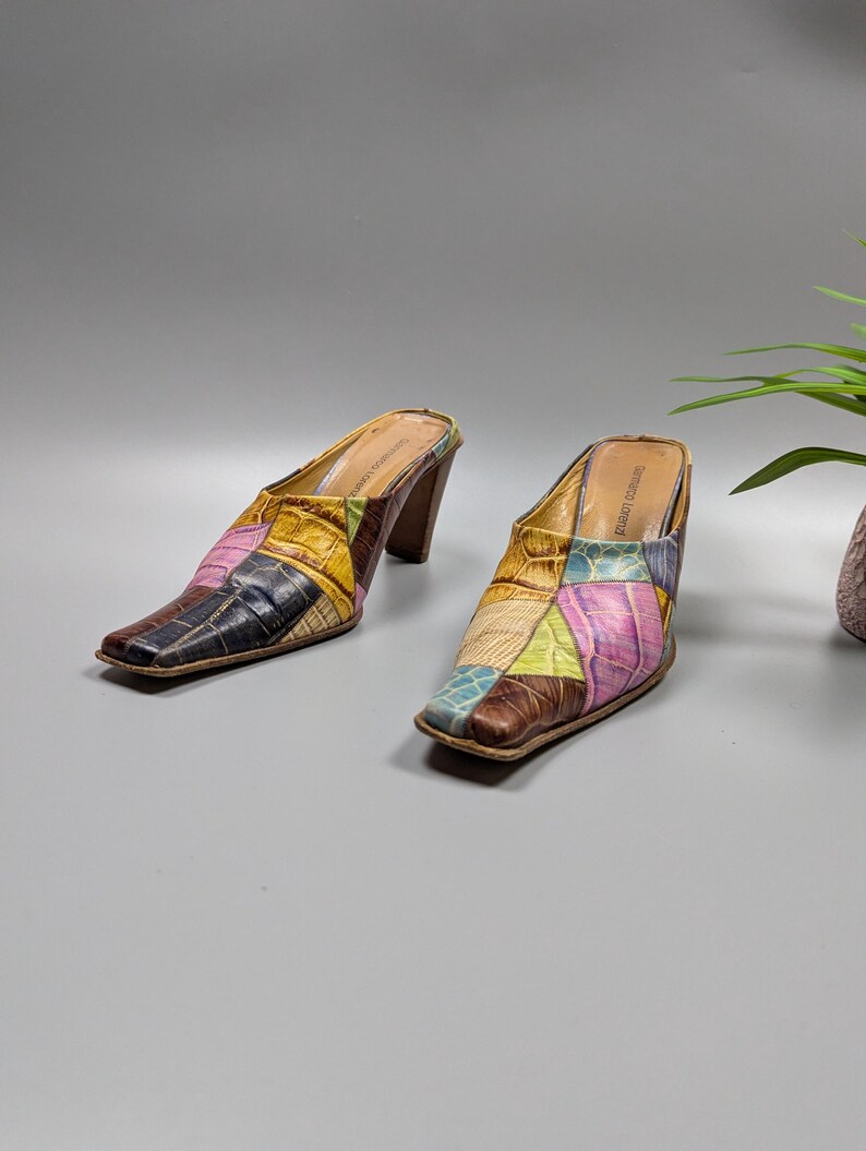 May include: A pair of patchwork leather mules with a square toe and a brown heel. The mules feature a variety of colors, including pink, blue, yellow, and brown. The shoes are set against a gray background.