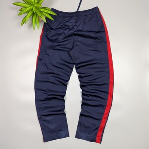 May include: Navy blue tracksuit bottoms with a red stripe down the side. The trousers have a drawstring at the waist. The trousers are made of a shiny material.