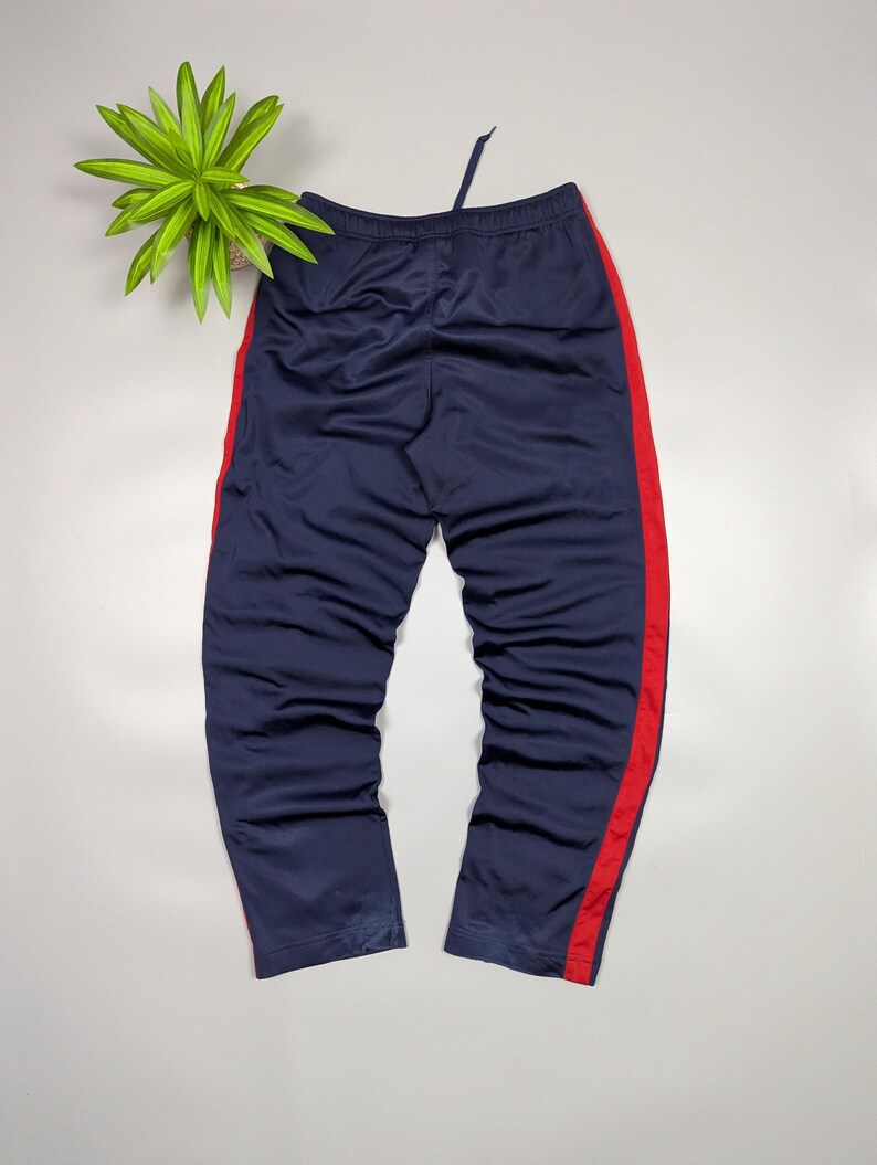 May include: Navy blue tracksuit bottoms with a red stripe down the side. The trousers have a drawstring at the waist. The trousers are made of a shiny material.