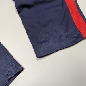 May include: Close-up of navy blue athletic trousers with a red stripe down the side. The fabric appears to be a smooth, synthetic material. The bottom hem of the trousers is frayed.