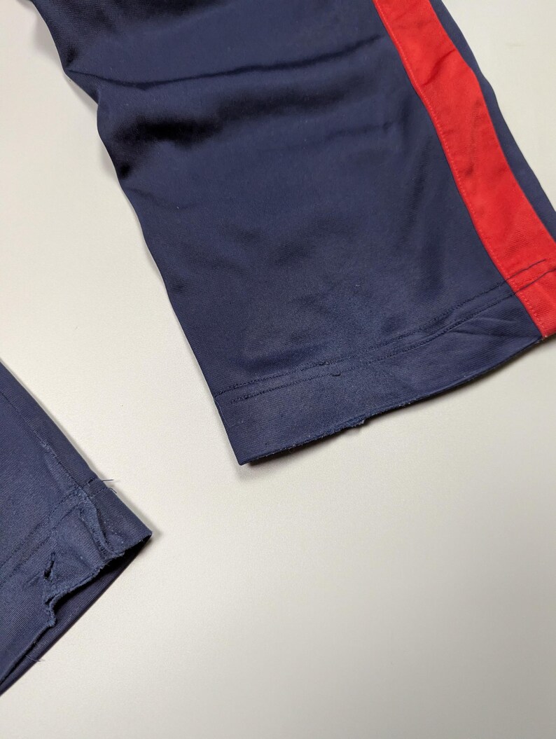 May include: Close-up of navy blue athletic trousers with a red stripe down the side. The fabric appears to be a smooth, synthetic material. The bottom hem of the trousers is frayed.