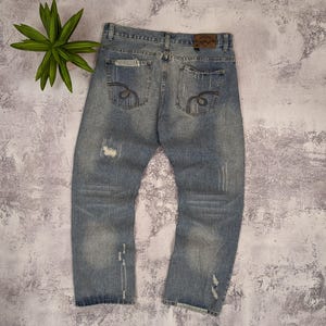 May include: Light blue denim jeans with distressed details, including rips and faded areas. The back pockets feature decorative stitching. A brown leather patch is visible at the waistband. The jeans are laid flat on a textured surface.