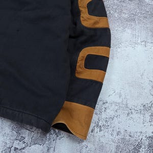 Pode incluir: Uma pe&ccedil;a de vestu&aacute;rio preta de manga comprida com cotoveleiras proeminentes em tom castanho-claro e um punho a condizer na extremidade da manga. O tecido parece ser de algod&atilde;o resistente ou lona com costuras vis&iacute;veis ao longo das bordas.