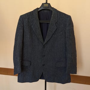 May include: A dark gray herringbone blazer with a three-button closure and two front pockets. The blazer is hanging on a black hanger against a neutral background. The interior lining is a dark navy blue.