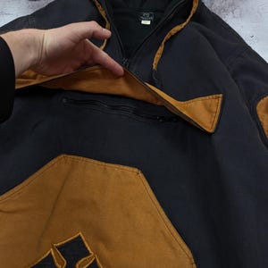 Pode incluir: Um casaco com capuz preto com detalhes em castanho, apresentando um bolso frontal grande com aba, um compartimento com fecho de correr e um emblema de cruz preta centrado na parte inferior do bolso. A pe&ccedil;a inclui capuz e mangas compridas.