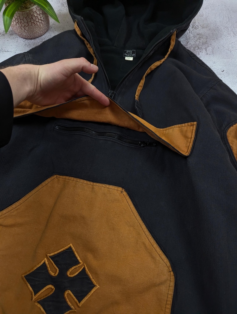 Pode incluir: Um casaco com capuz preto com detalhes em castanho, apresentando um bolso frontal grande com aba, um compartimento com fecho de correr e um emblema de cruz preta centrado na parte inferior do bolso. A pe&ccedil;a inclui capuz e mangas compridas.