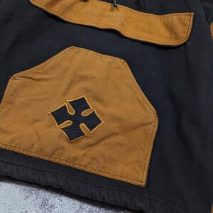 Pode incluir: Um moletom com capuz preto e castanho com um grande bolso canguru frontal apresentando um emblema de cruz preta. A pe&ccedil;a inclui um decote com fecho de correr parcial, capuz com cord&atilde;o e mangas compridas com pain&eacute;is castanhos nos ombros.