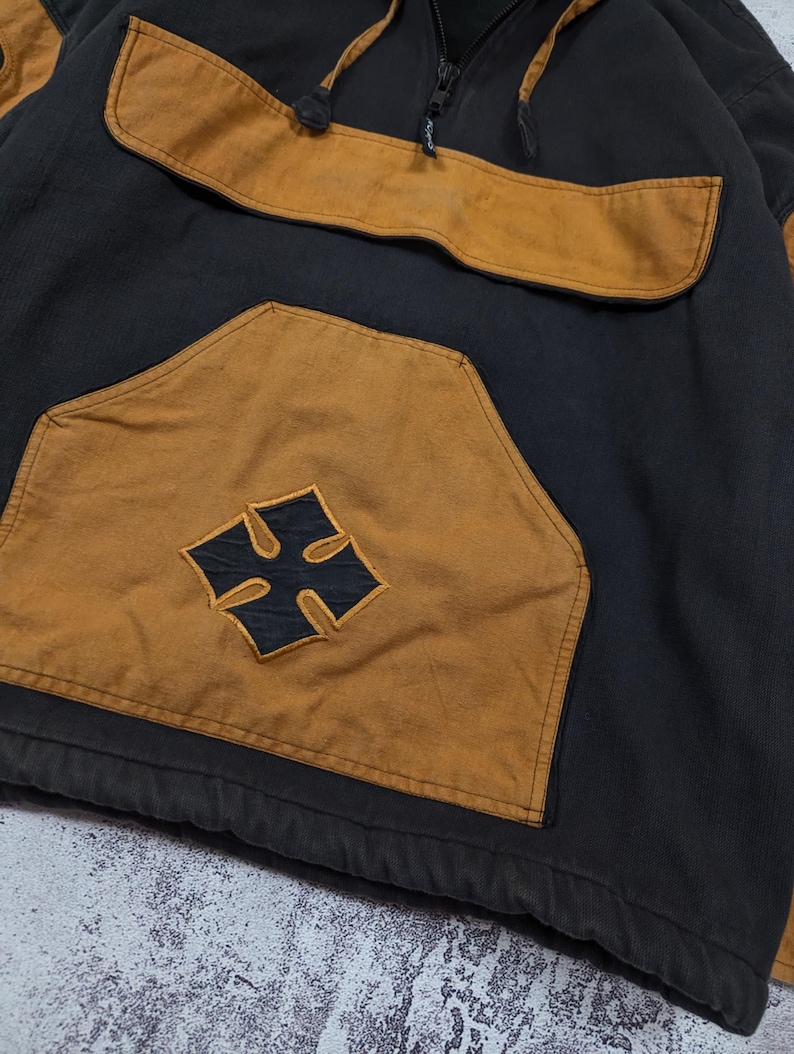 Pode incluir: Um moletom com capuz preto e castanho com um grande bolso canguru frontal apresentando um emblema de cruz preta. A pe&ccedil;a inclui um decote com fecho de correr parcial, capuz com cord&atilde;o e mangas compridas com pain&eacute;is castanhos nos ombros.
