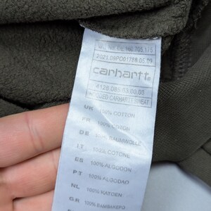 Puede incluir: Primer plano de una etiqueta Carhartt en una sudadera con capucha verde oscuro. La etiqueta incluye el logotipo de Carhartt, informaci&oacute;n del producto y detalles de la composici&oacute;n de la tela en varios idiomas, incluido 100% algod&oacute;n.
