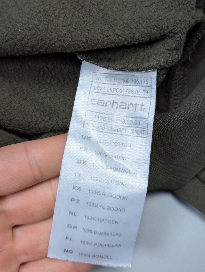 Puede incluir: Primer plano de una etiqueta Carhartt en una sudadera con capucha verde oscuro. La etiqueta incluye el logotipo de Carhartt, informaci&oacute;n del producto y detalles de la composici&oacute;n de la tela en varios idiomas, incluido 100% algod&oacute;n.