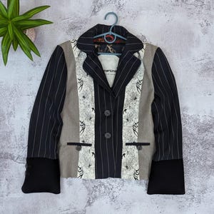 Könnte beinhalten: Ein Blazer aus einer Kombination von schwarzem Nadelstreifenstoff, beigen Nadelstreifenbahnen und weißen floralen Mustereinsätzen. Das Kleidungsstück hat ein steigendes Revers, zwei Frontknöpfe, zwei Leistentaschen und breite schwarze Ärmelbündchen.