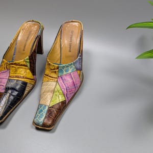 May include: A pair of patchwork leather mules with a square toe and a stacked heel. The shoes feature a variety of colors, including yellow, blue, pink, and brown. The brand name "Gennaro Lorenzi" is visible on the inner sole.